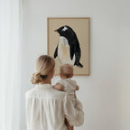 Woman holding a baby looking at a framed penguin artwork on a wall.